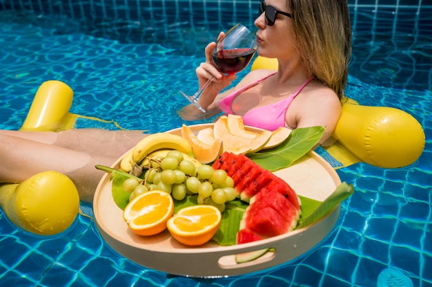 Photo beautiful woman lying on floating hammock in the swimming pool with wine and tray of fruits