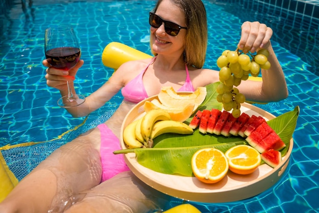 Photo beautiful woman lying on floating hammock in the swimming pool with wine and tray of fruits