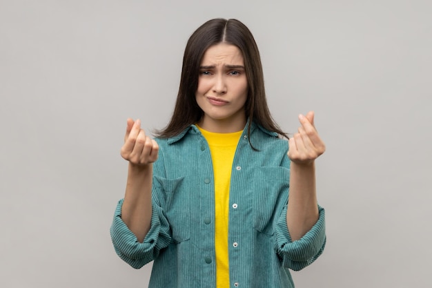 Beautiful woman makes money gesture rubs fingers looking at camera with glad expression