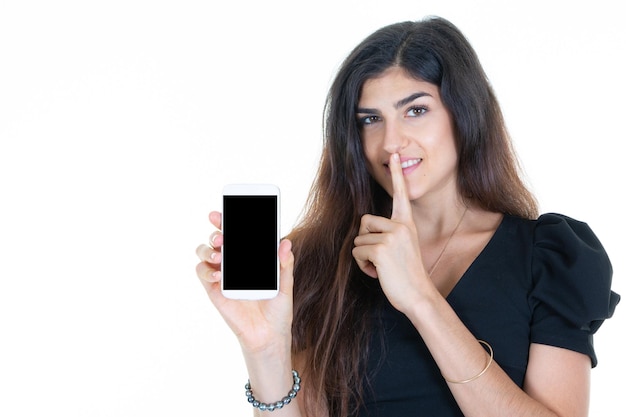 Beautiful woman makes secret silence gesture on lips while holding smartphone which has empty black screen of phone on white background
