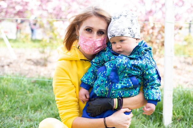 Beautiful woman in medical mask with little baby boy near pink sakura blooming tree Spring concept