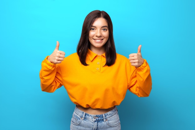 Beautiful woman of mixed ethnicity shows likes with thumbs in orange shirt against blue background