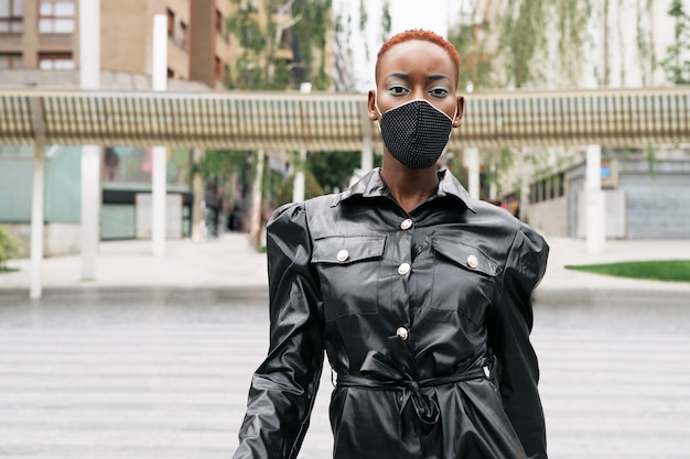 Красивая женщина-модель с маской из-за пандемии коронавируса covid 19 стильно гуляет по улице в красивом черном платье