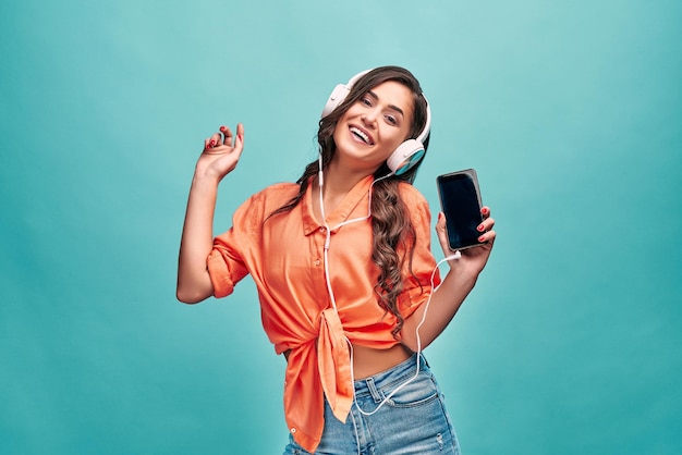 Красивая женщина в оранжевой рубашке танцует, смеется, показывает телефон на камеру и слушает музыку