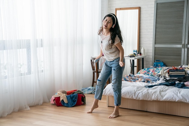 Beautiful woman packing suitcase in cozy bedroom. full length happy young asian female standing on wooden floor hold mobile phone with headphones dancing in modern house. cheerful funny girl pose