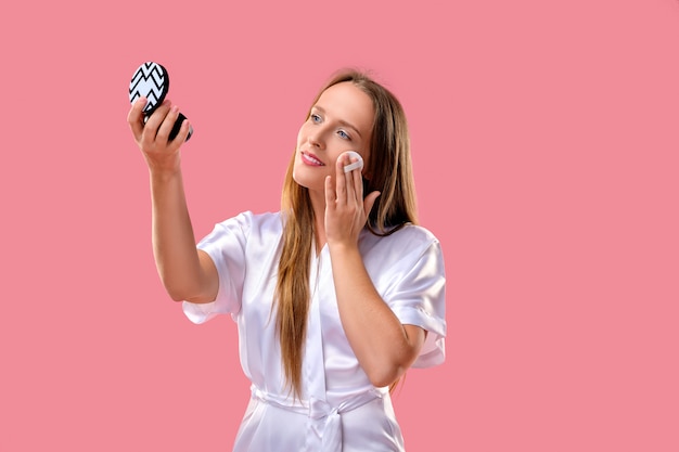 Beautiful woman on pink background applying foundation on her face