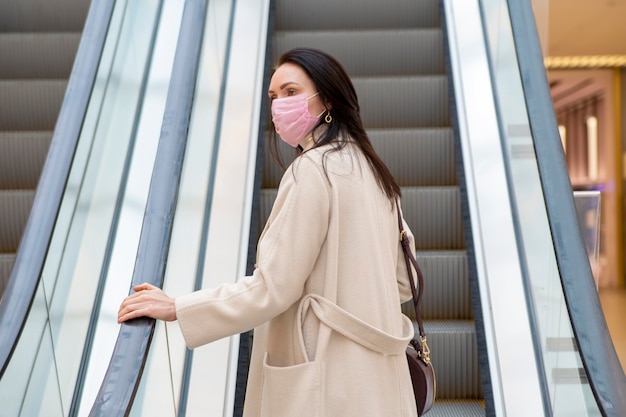 beautiful woman in a pink medical mask on her face against the background of the escadator in a public place.