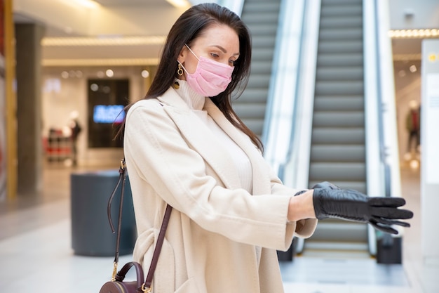 beautiful woman in pink medical mask putting on leather gloves in a public place on background escalator