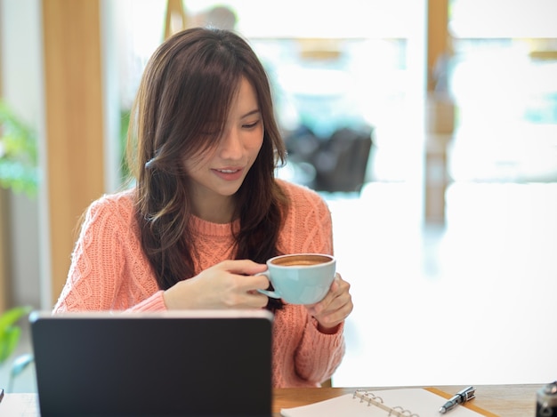 분홍색 스웨터를 입은 아름다운 여성이 태블릿에서 온라인으로 공부하는 동안 카페에서 커피를 마신다