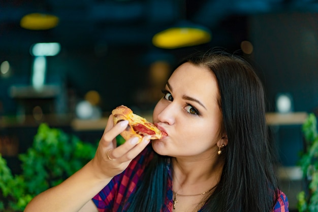 A beautiful woman in a plaid shirt holds a slice of pizza in her hand and eats it in a pizzeria. 