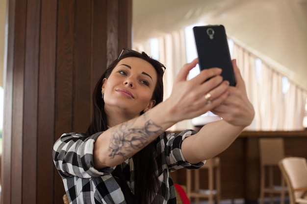 Beautiful woman and plaid shirt is doing selfie on a black phone in a beautiful modern cafe