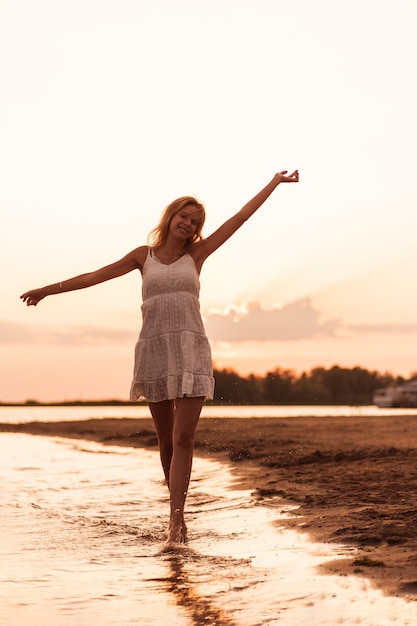 Beautiful woman posing on the beach a slender young blonde in a white dress is spinning on the river...