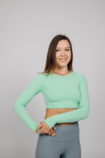 Beautiful woman posing in studio Portrait of happy young brunette in green top for yoga or fitness she stands on gray isolated background and smiles shows off her athletic figure