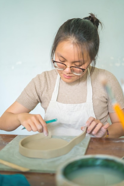 Beautiful woman potter working on potters wheel making ceramic\
pot from clay in pottery workshop focus hand young woman attaching\
clay product part to future ceramic product pottery workshop