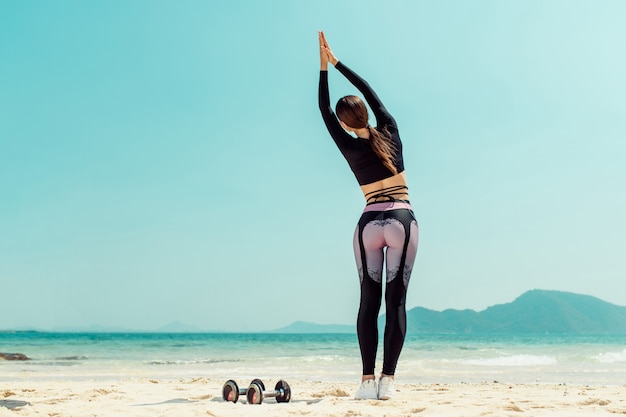 Beautiful woman practices yoga by the sea on a sunny day. The woman does stretching exercises. Dumbbells lying sand. Back view