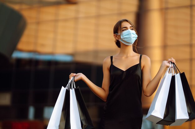 Beautiful woman in protective sterile medical mask with shopping bags near shopping center.