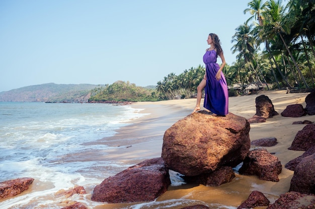 Foto bella donna in un abito lungo viola in piedi su una pietra in riva al mare sullo sfondo di palme cola colva palolem beach a goa india copyspase