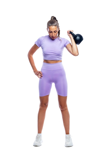 A beautiful woman in purple sportswear is doing exercises with a kettlebell Sport health and activity Isolated on white background Full height Vertical