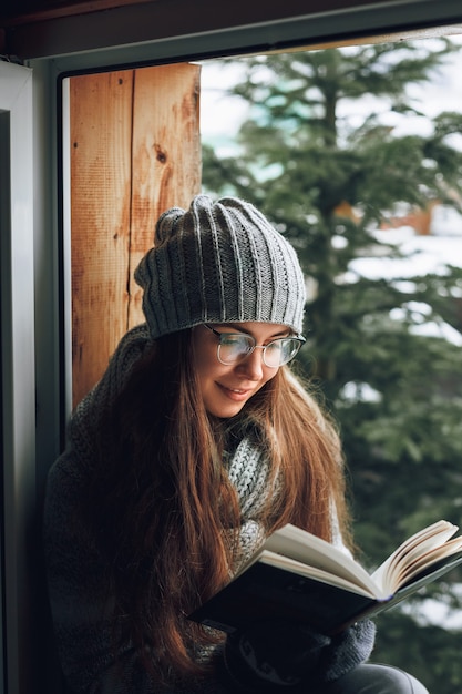 Beautiful woman reading a book in a sweater  sitting home by the window