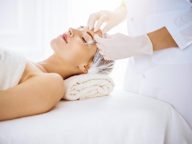 Beautiful woman receiving beauty injections with closed eyes in sunny medical center. Beautician doctor hands doing beauty procedure to female face with syringe. Cosmetic medicine and surgery concept.