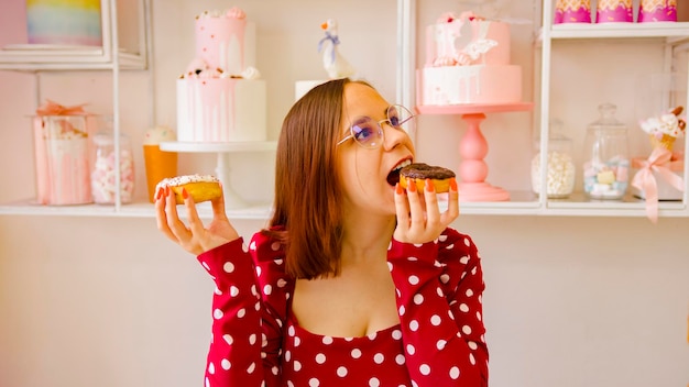 A beautiful woman in a red dress and glasses holds a white donut in one hand and bites a chocolate donut while holding it with the other hand in the cafe