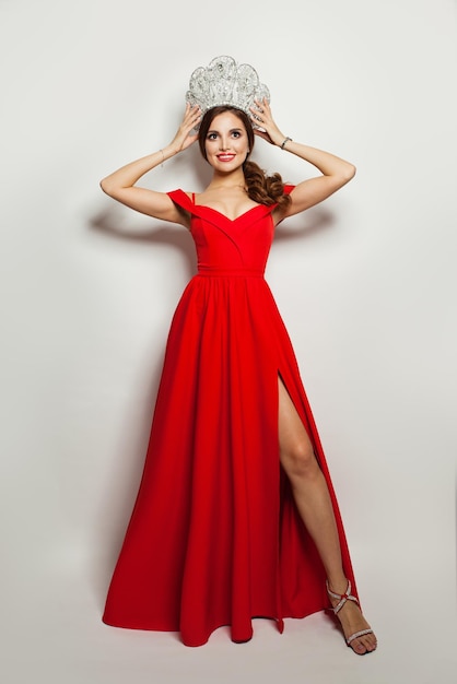 Beautiful woman in red dress puts on diamond crown against white wall background