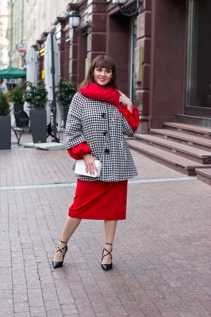 A beautiful woman in a red dress and scarf. Smiling and shy in a hat on the square in the city