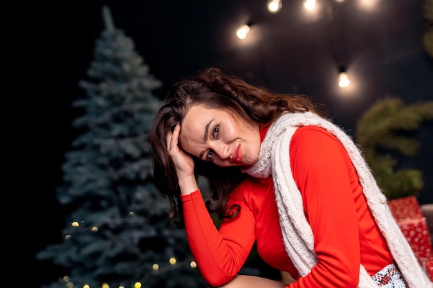 Beautiful woman in a red sweater and white scarf sitting with the hand at her face Pretty girl with festive lights and Christmas tree at the background