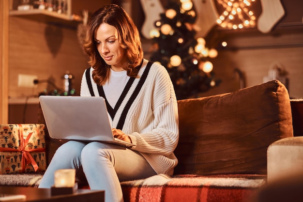 Bella donna che si rilassa sul divano facendo acquisti di natale su internet nella stanza decorata nel periodo natalizio.