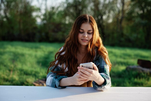 A beautiful woman relaxing and working on her phone sitting in nature in the park among the trees smiling and holding her smartphone in her hand lit by the bright sunset light High quality photo