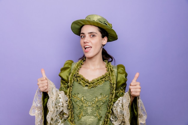 Beautiful woman in renaissance dress and hat happy and positive smiling cheerfully showing thumbs up on blue