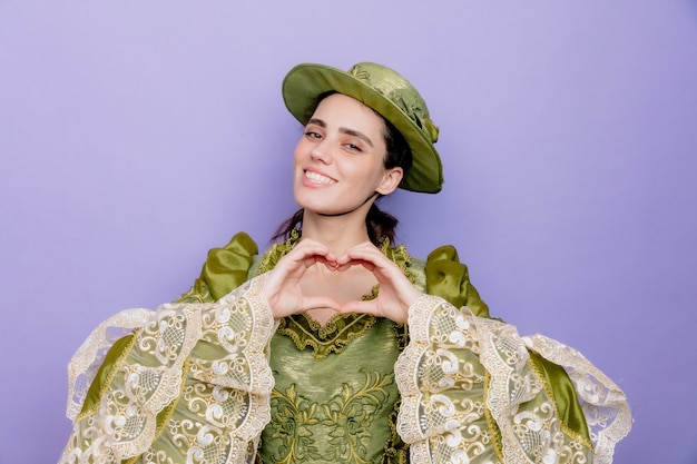 Beautiful woman in renaissance dress and hat smiling friendly making heart gesture with fingers happy and positive on blue