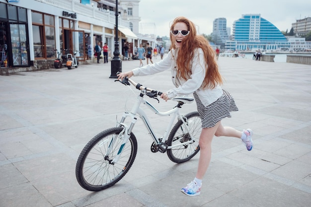 Beautiful woman riding on bike Lifestyle and health in the city Cheerful redhaired young woman gets pleasure from walking around the city