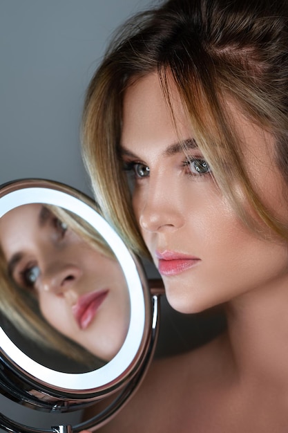 Beautiful woman and round mirror with LED light on gray background