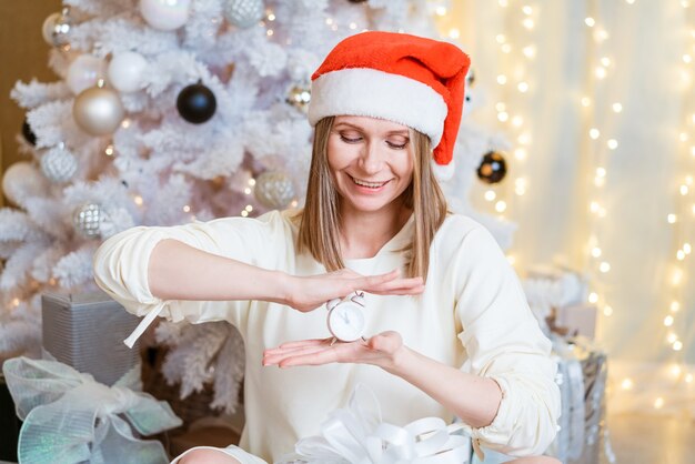 산타 모자를 쓴 아름다운 여성이 알람 시계를 들고 축제 크리스마스 배경에 귀여운 미소를 짓고 있습니다.