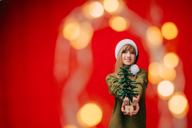 A beautiful woman in a Santa hat holds a small Christmas tree in her hands New year concept