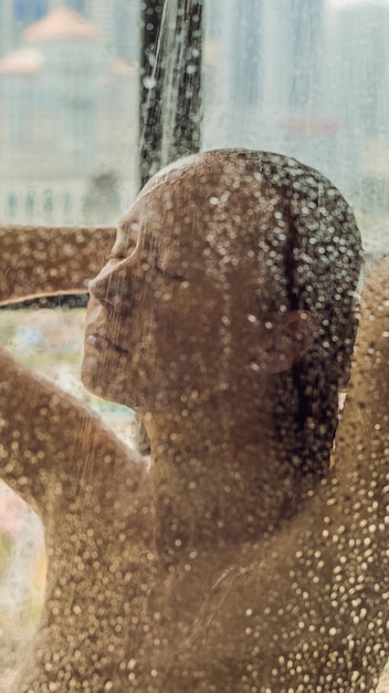 탁 트인 창문 배경에 물방울이 있는 유리 뒤 샤워에 있는 아름다운 여성