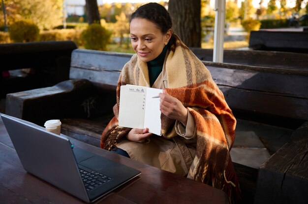 Bella donna che mostra una pagina vuota del quaderno durante la videochiamata sul laptop, si tiene al caldo avvolgendosi in una coperta di lana, seduta all'aperto su una panca di legno. lavoro a distanza, concetto di apprendimento su internet