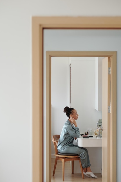 Beautiful woman in silk pajamas sitting in front of vanity and applying skincare and makeup