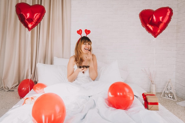 Красивая женщина сидит в постели с белым постельным бельем и ест шоколад. День святого Валентина.