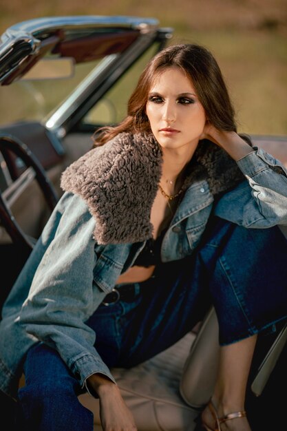 a beautiful woman sits on a convertible in a field dressed in jeans and a fur jacket beautiful and stylish look