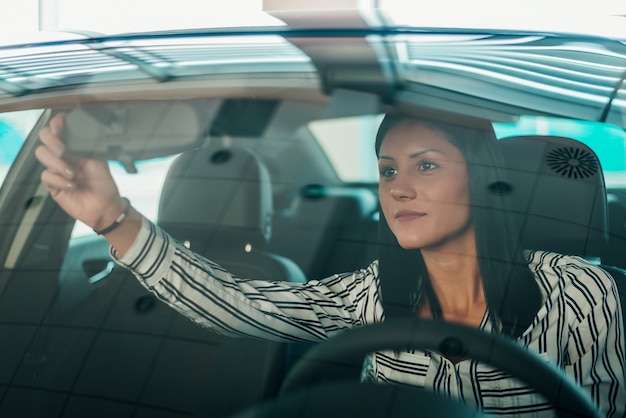 美しい女性が車の中で座って、車のミラーを調整します。