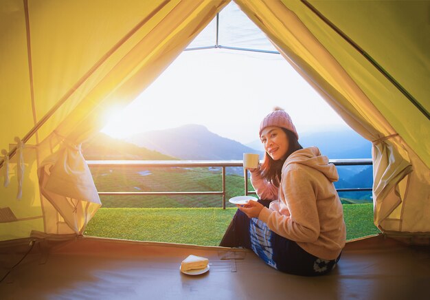 A beautiful woman sitting and drinking coffee in a morning tent with a mountain 