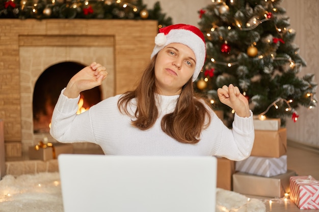 Beautiful woman sitting on floor working with laptop at home around christmas tree, stretching her back, tired and relaxed, girl wearing white sweater and santa claus hat