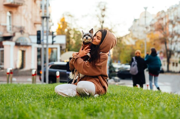 通りの背景に彼女の腕の中でかわいい小さな犬と一緒に芝生に座っている美しい女性