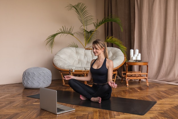 Beautiful woman sitting in lotus position and doing breathing exercises watching workout on laptop