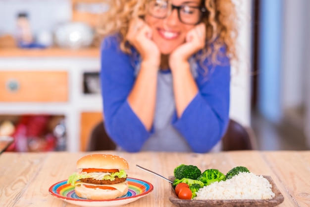 Beautiful woman smiling and looking at the table - choosing hamburger or rise and vegetables - sane and health lifestyle concept