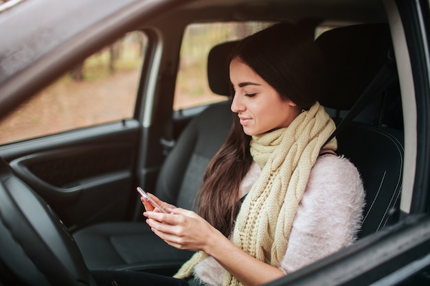 Bella donna che sorride mentre seduto in macchina. la ragazza sta usando uno smartphone.