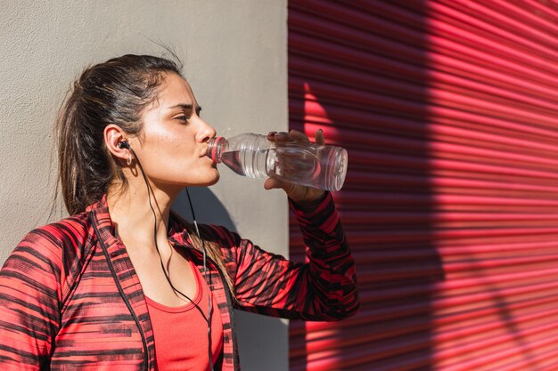 Beautiful woman in sportswear drinks water from a bottle at sunset in the city