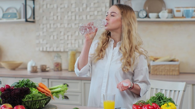 Beautiful woman standing in the kitchen and drinks mineral sparkling water from a plastic bottle